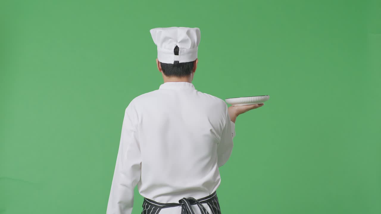 Vista posteriore di uno chef asiatico che tiene un piatto mentre cammina in uno studio con sfondo green screen