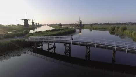 Un-Dron-Disparó-A-La-Izquierda,-Alrededor-De-Una-Niña-Caminando-Y-Dando-La-Vuelta-En-Un-Puente,-Mirando-Los-Molinos-De-Viento-Holandeses-En-Los-Países-Bajos-Durante-El-Amanecer