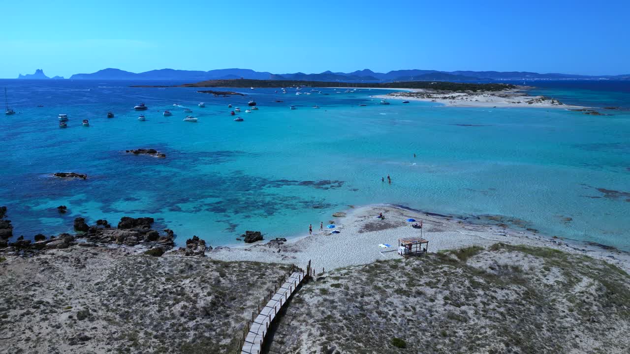 Espalmador island featuring white sand beaches, clear turquoise water, and anchored boats. Ibiza and the island of Es Vedra in the background. Lovely aerial view flight descending drone
