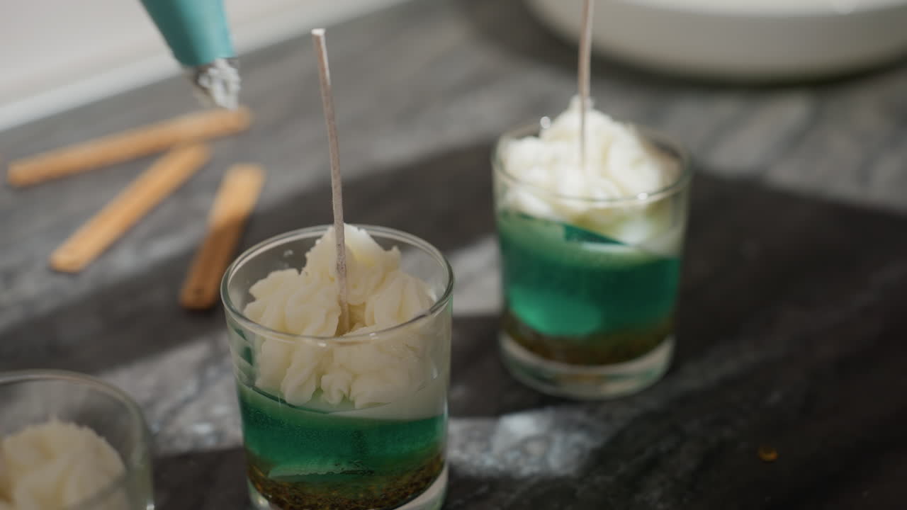 Culinary expert creates elegant milk-colored floral patterns on dessert cups using piping bag, forming intricate swirls and peaks in clear containers filled with green layered base