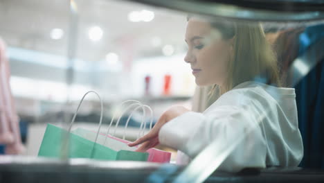 mujer joven sentada, vista a través del vidrio, abriendo suavemente la bolsa de compras de menta en una moderna tienda minorista, rodeada de iluminación ambiental y colores vibrantes