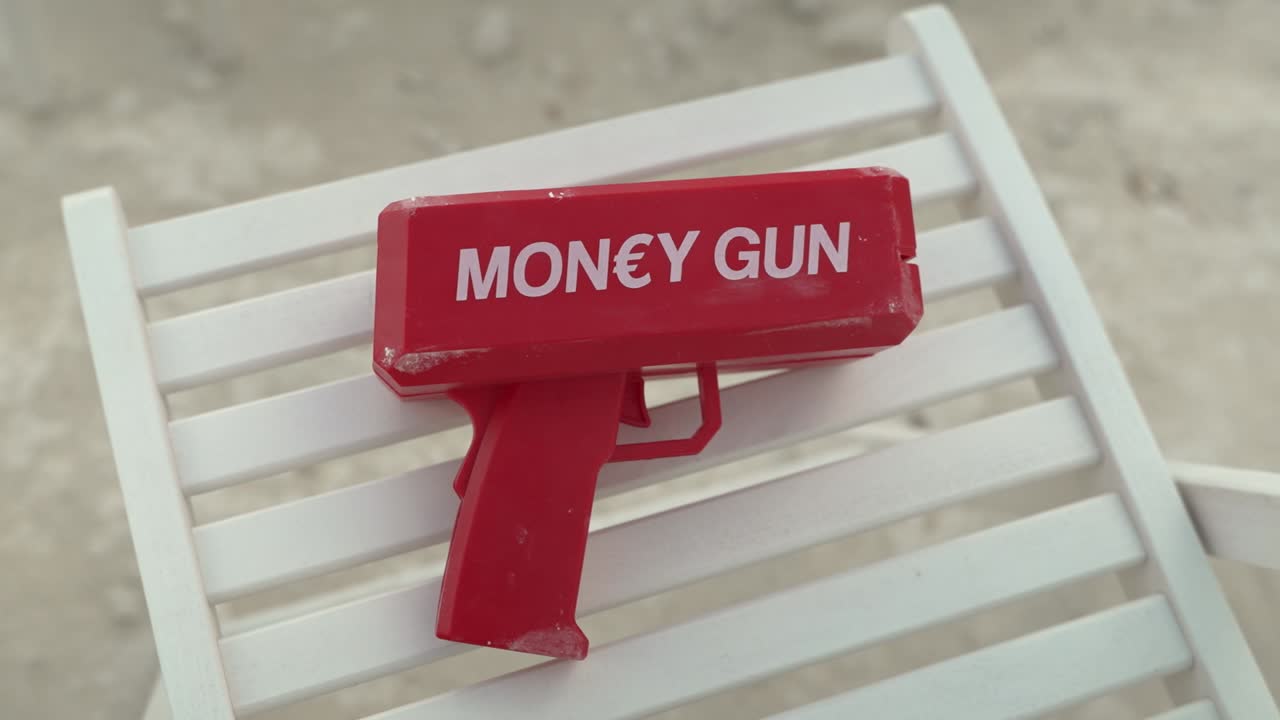 A red money gun rests on a white beach chair. Sunbathers lounge under the sun while children play in the sand. The lively beach atmosphere enhances the forgotten toy's presence