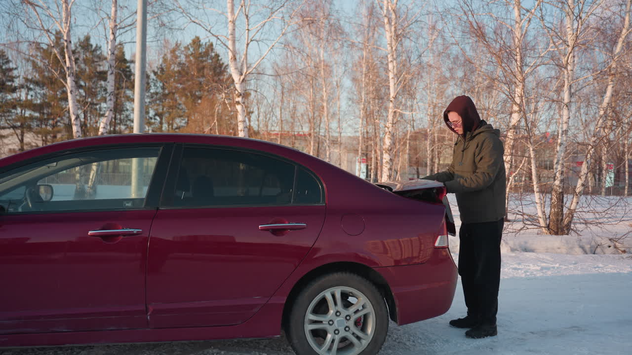 Man in hoodie shuts trunk of red car and walks backward in snowy lot with bare trees in background and faint outline of another car visible through branches