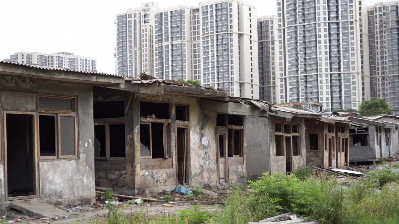 Edifici a un piano logori passano attraverso l'inquadratura durante una lenta panoramica a sinistra, rivelando porte aperte, intonaco scrostato, danni al tetto e detriti sotto luminose torri residenziali.