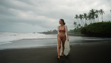 Smiling,-young-model-in-red-bikini-walking-on-black-sand-beach-in-tropical-country