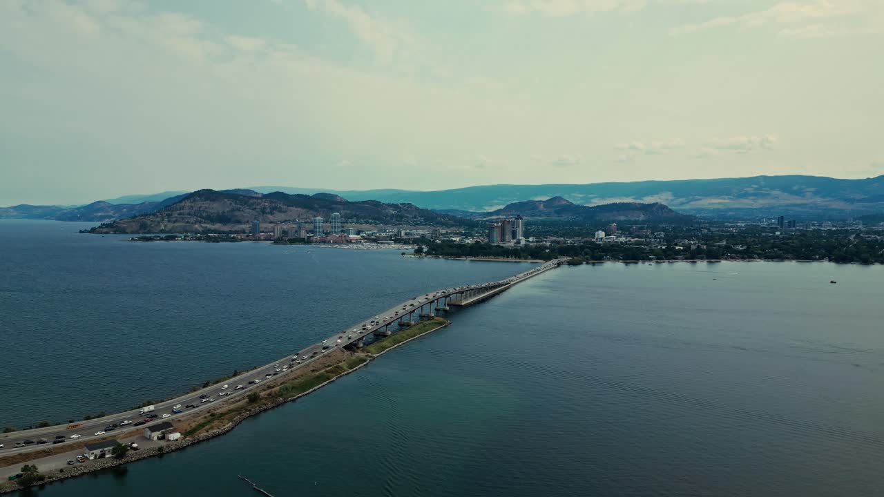 High flying Aerial over Okanagan Lake view William R Bennett Bridge Central Okanagan BC calm blue water traffic cars trucks commuting over water mountainous background light blue sky wispy clouds