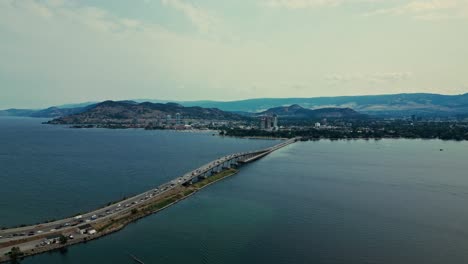 high flying aerial over okanagan lake view william r bennett bridge central okanagan bc calm blue water traffic cars trucks commuting over water mountainous background light blue sky wispy clouds
