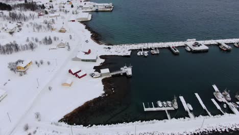 Drone-view-in-Tromso-area-in-winter-flying-over-a-snowy-landscape-surrounded-by-the-sea-and-a-frozen-port-with-boats-and-white-mountains-in-Norway