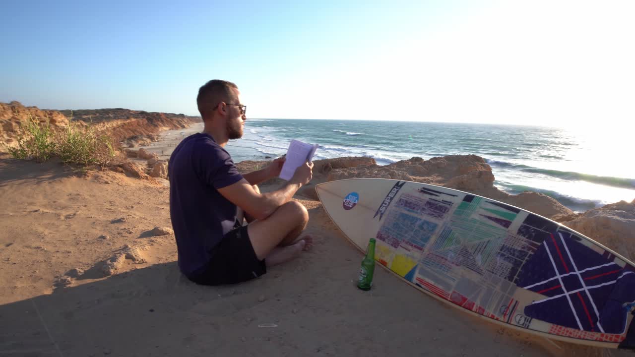 toma cinematográfica giratoria de una persona leyendo un libro sentada cerca de una tabla de surf y una botella de cerveza en una playa en hadera, israel