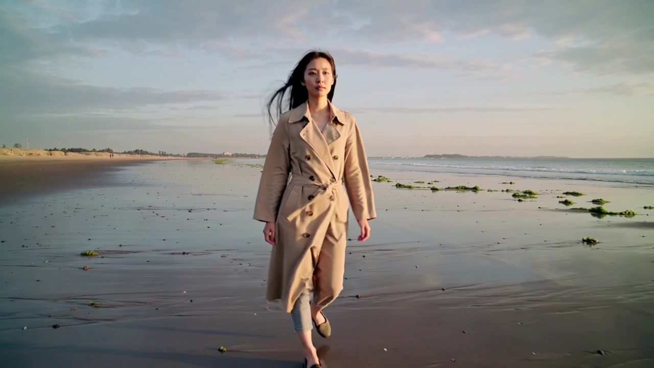 Starting from afar, woman in beige trench coat walking purposefully toward camera across wet sand