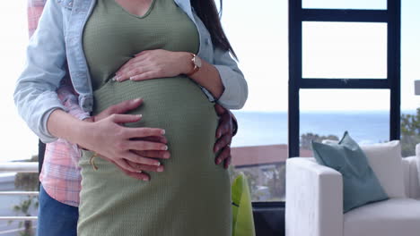 expecting couple embracing, hands on pregnant belly, standing in living room, copy space
