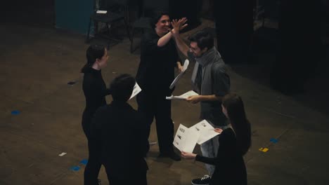 A-happy-and-cheerful-group-of-actors-in-black-suits-together-with-their-stage-director-in-a-gray-T-shirt-and-scarf-are-holding-sheets-of-paper-with-a-script-in-their-hands-and-are-ready-to-go-on-stage-with-the-presentation-of-their-play-in-the-theater