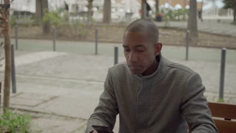 joven con abrigo usando un teléfono inteligente y bebiendo de una taza de papel