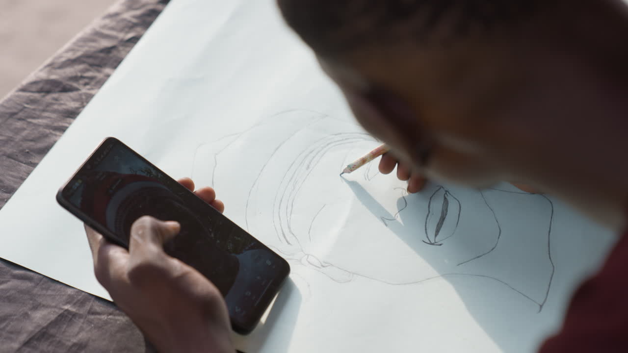 芸術家が熱心にスケッチする。精巧なスマートフォンの絵を描くイラストレーター。熟練した黒人イラストレーターが、ガイドに沿ってスマートフォンの参考資料を正確な集中力と意図をもって注意深くスケッチする。