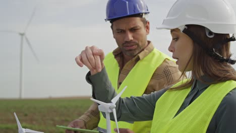 Latin-man-and-caucasian-woman-professionals-standing-on-wind-turbine-field-and-discussing.