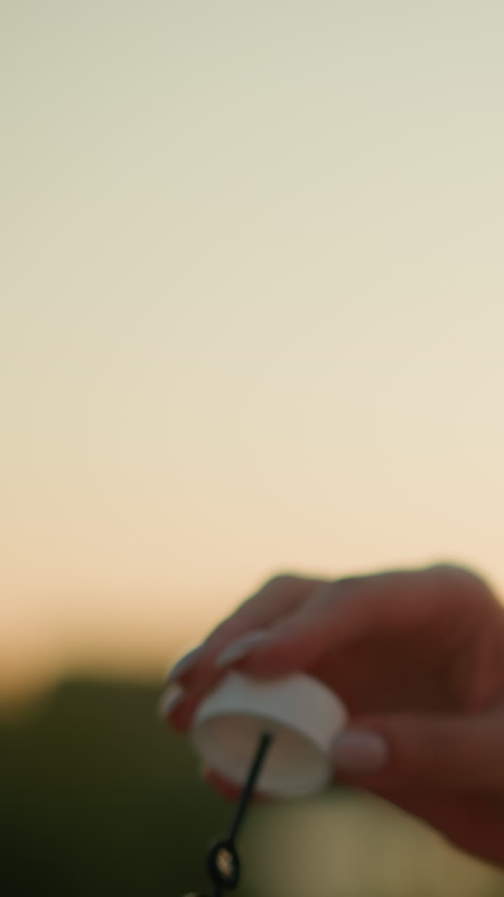 primer plano de una mujer blanca soplando burbujas al aire libre durante la puesta del sol con gafas de sol en la cabeza, la luz del sol ilumina suavemente su cara mientras sostiene la varita de burbujas