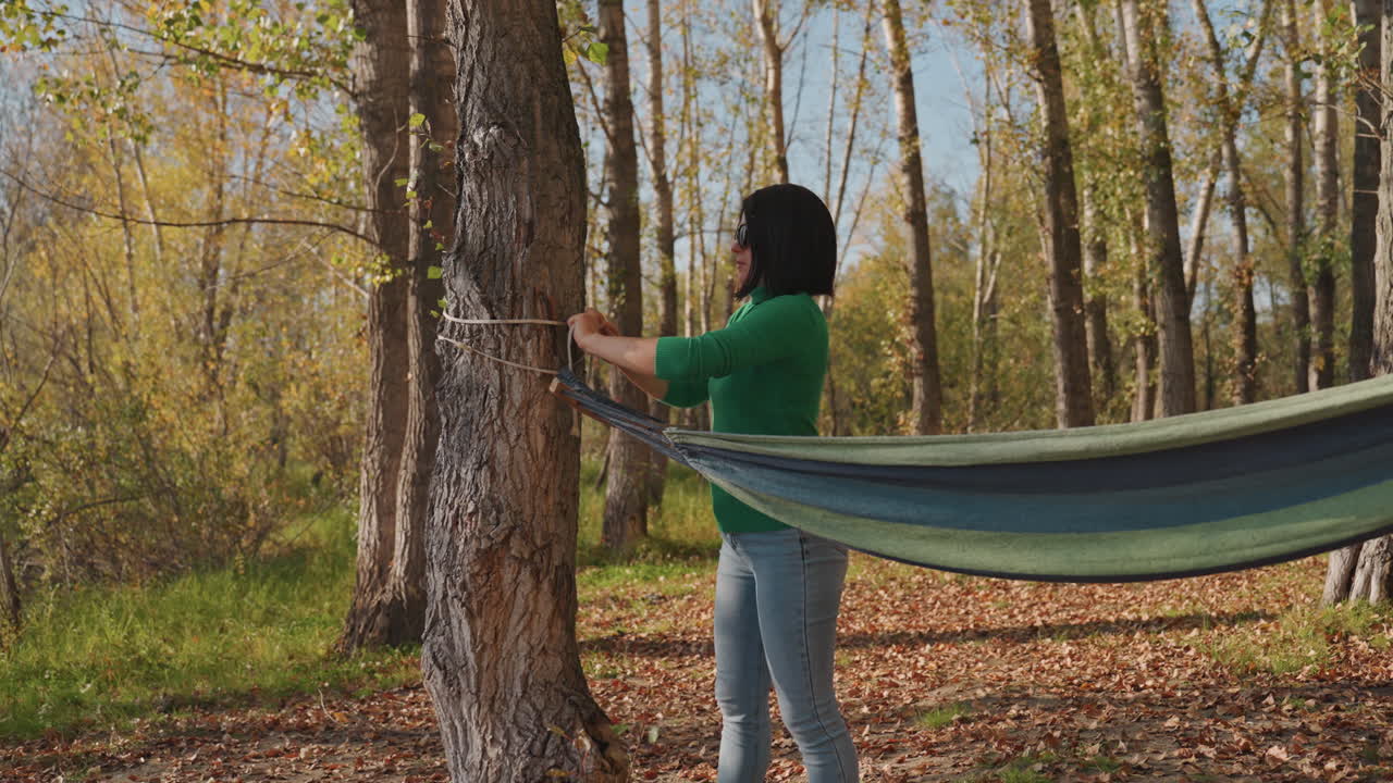Donna che fissa un'amaca a un albero, facendo passare la corda attraverso un moschettone e stringendo la cinghia, testando il punto di ancoraggio in un bosco luminoso, primo piano di mani che annodano, preparazione organizzata del campeggio all'aperto.