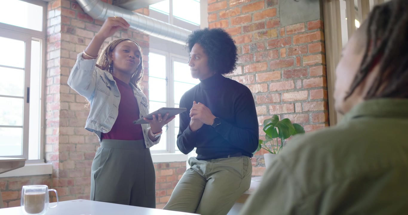 Colegas de trabalho afro-americanas fazendo uma apresentação na mesa, segurando um tablet e gesticulando perto de um laptop e uma planta.