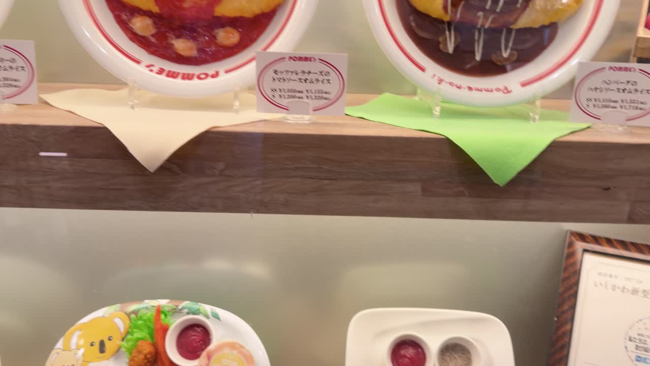 Una telecamera a mano esegue una panoramica su dettagliate repliche di cibo in plastica all'interno di una vetrina di un ristorante di Kanazawa