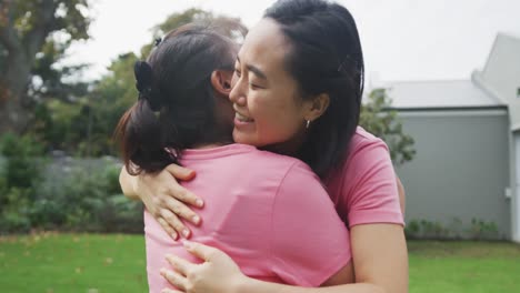 happy asian mother and adult daughter embracing and high fiving in garden, wearing pink t shirts