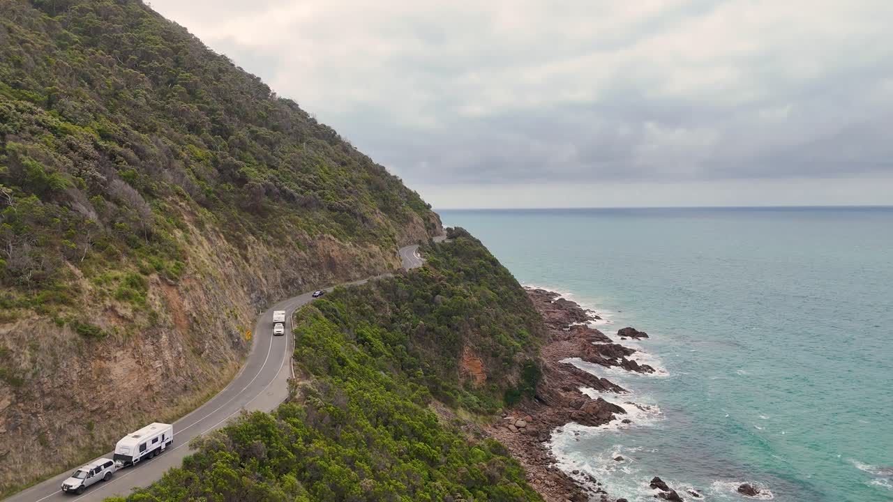 Aerial view of vehicles navigating a coastal road beside lush cliffs and turquoise ocean under cloudy skies