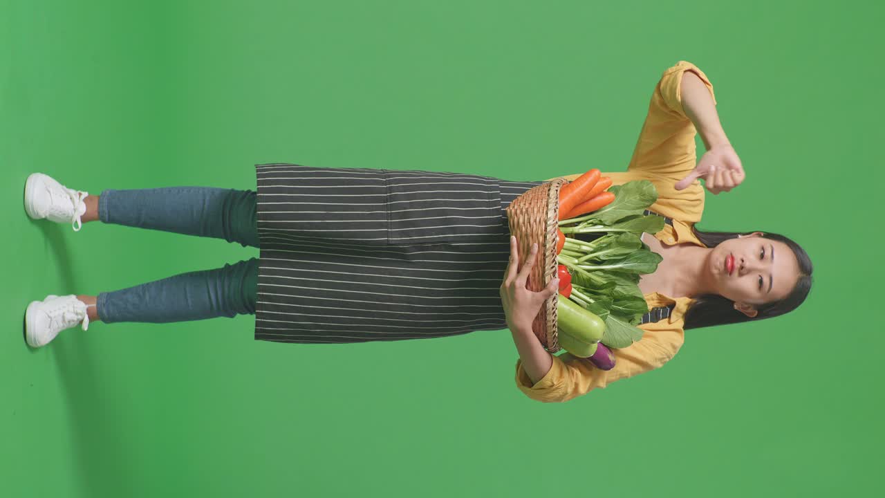 cuerpo lleno de agricultora asiática con canasta de verduras que muestra los pulgares hacia abajo gesto mientras está de pie en el estudio de pantalla verde