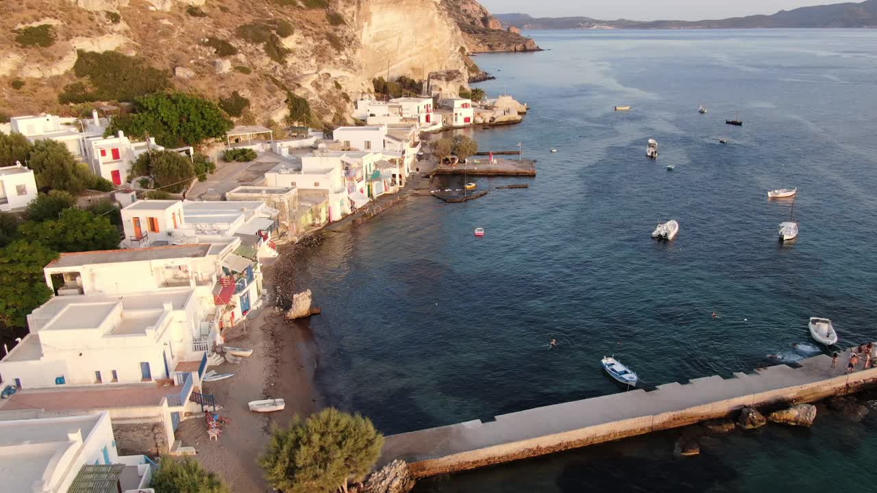 vista de dron en grecia volando sobre una pequeña ciudad con casas blancas y puertas de colores al lado del mar y la montaña en milos al atardecer