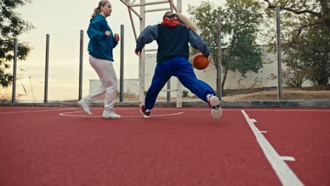 Filmada-Desde-Abajo,-Una-Chica-Rubia-Con-Pantalones-Azules-Y-Uniforme-Deportivo-Corre-Por-La-Cancha-Roja-Y-Acusa-A-Todos-Los-Oponentes-De-Lanzar-Una-Pelota-Naranja-A-Un-Aro-De-Baloncesto-En-Verano.