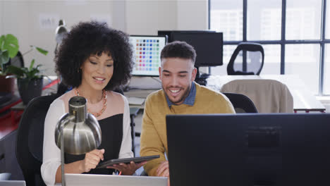 happy diverse male and female colleague at desk, using tablet, computer and talking, slow motion