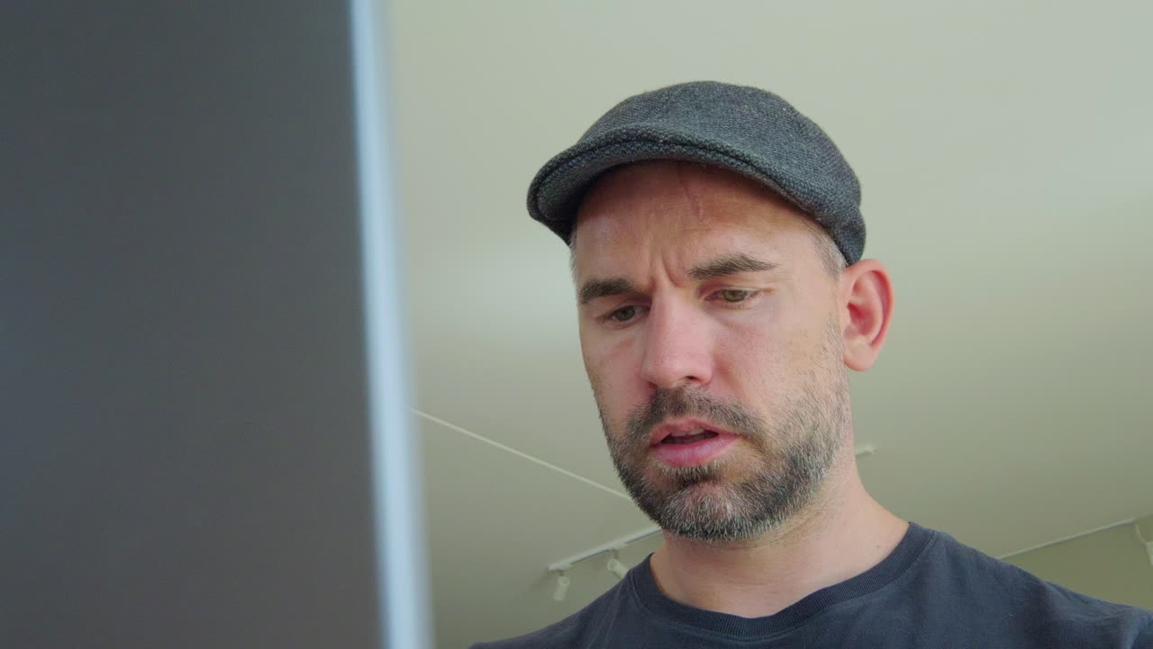 Serious Man In Flat Cap Typing On A Laptop Computer At Home