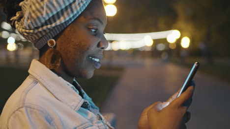 Lächelnde-Afroamerikanische-Frau,-Die-Abends-Draußen-Auf-Dem-Smartphone-Tippt