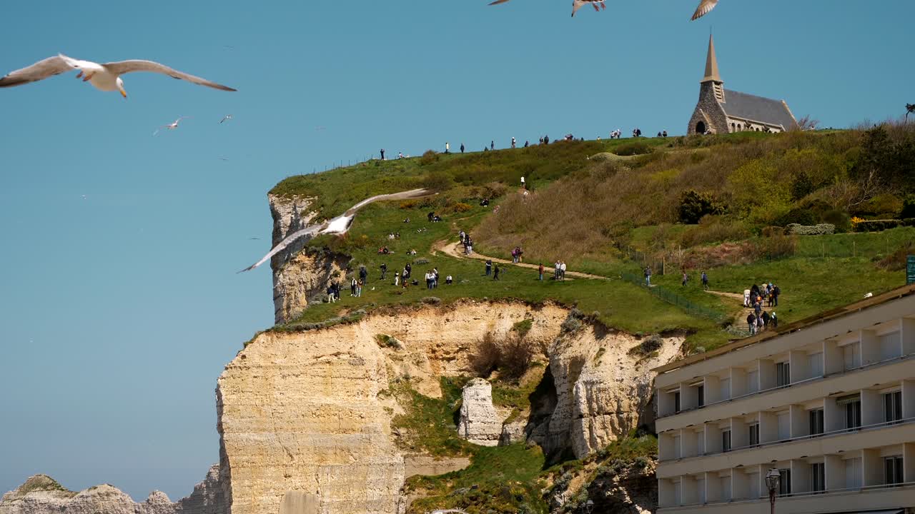 Slow Motion shot of seagulls flying over coast of Etretat with famous church on hilltop and golden sea cliffs during golden sunset - France,Europe - Wide shot