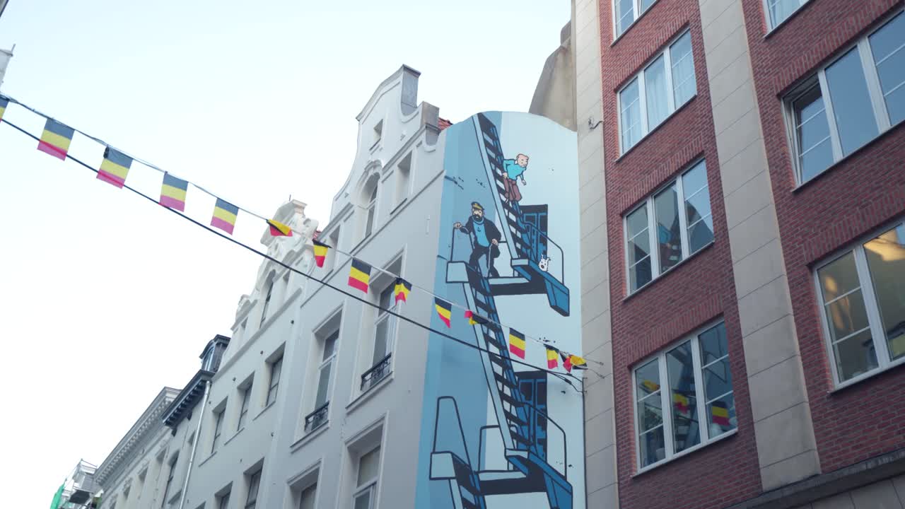 Tintin and Captain Haddock Mural, Brussels, Belgium, small belgian flags hanging, waving across street. Panning rotational shot. Clear skies. Cartoon painting on old mansion house wall. City center.