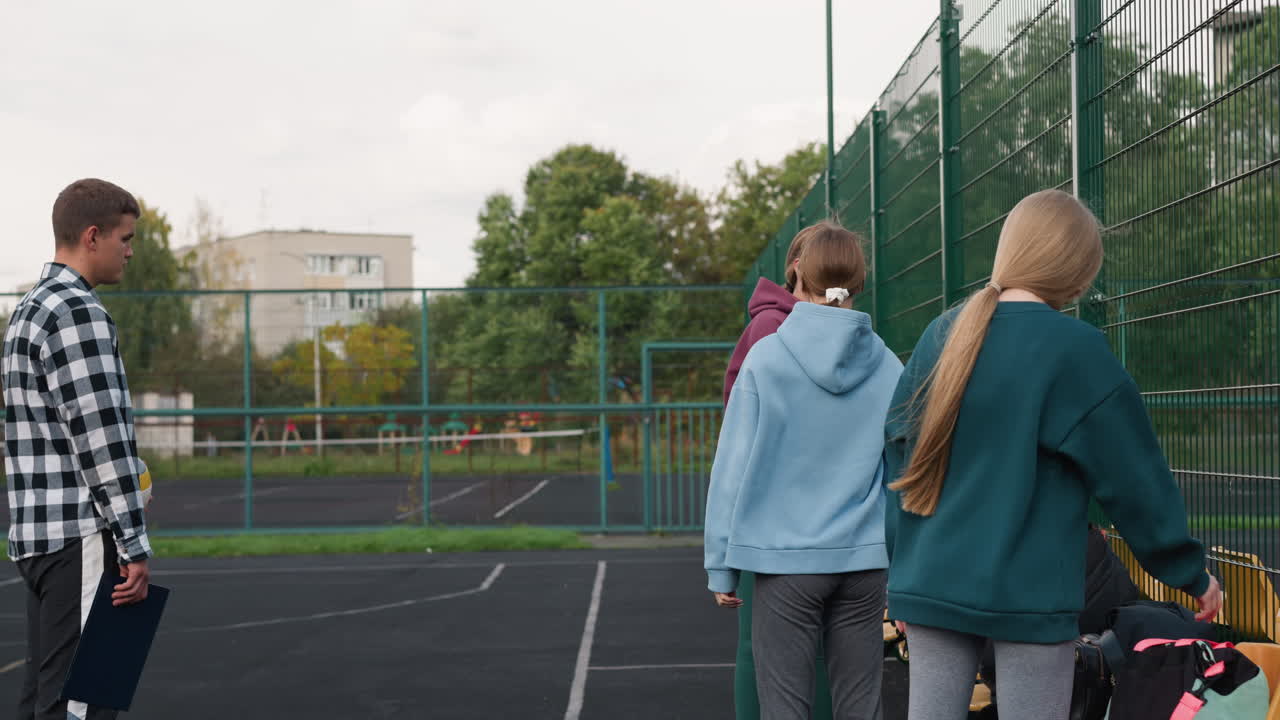 학생들이 배구장, 나무, 주거 건물 및 스포츠 시설의 벤치에 가방을 놓을 때 기다리는 동안 성능 책을 들고 배구 공을 뛰는 코치