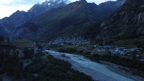 una foto de un dron de un pequeño y pintoresco pueblo remoto a orillas del río baspa con los picos nevados del himalaya indio al fondo