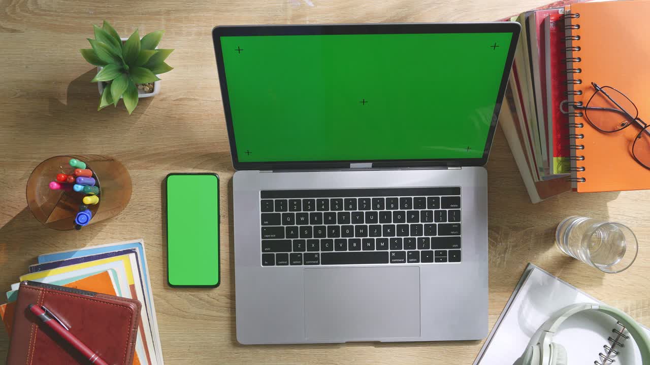 Top down view of a laptop computer with mock up green screen chromakey display on a wooden office desk next to notebook with pens, glasses, and a glass of water. Slow zoom out, close up