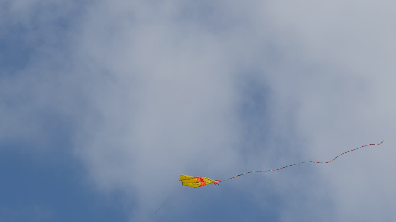 A yellow kite with a vibrant tail flies high in a cloudy blue sky on a windy day