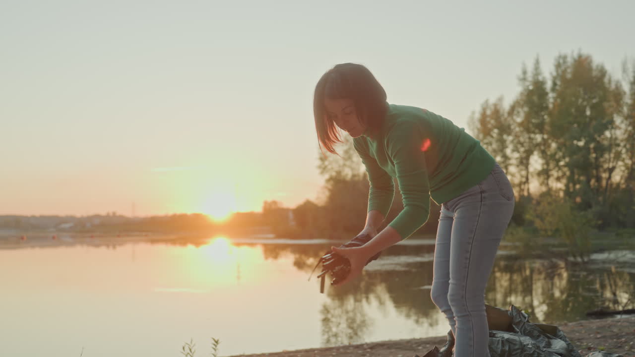Kvinde campingplads solnedgang. Rolig ramme viser kvinde der organiserer udstyr, mens solen går ned bag vandet. Fredfyldt billede af kvinde der forbereder sin campingplads med solnedgangens glød på den blide vandoverflade.