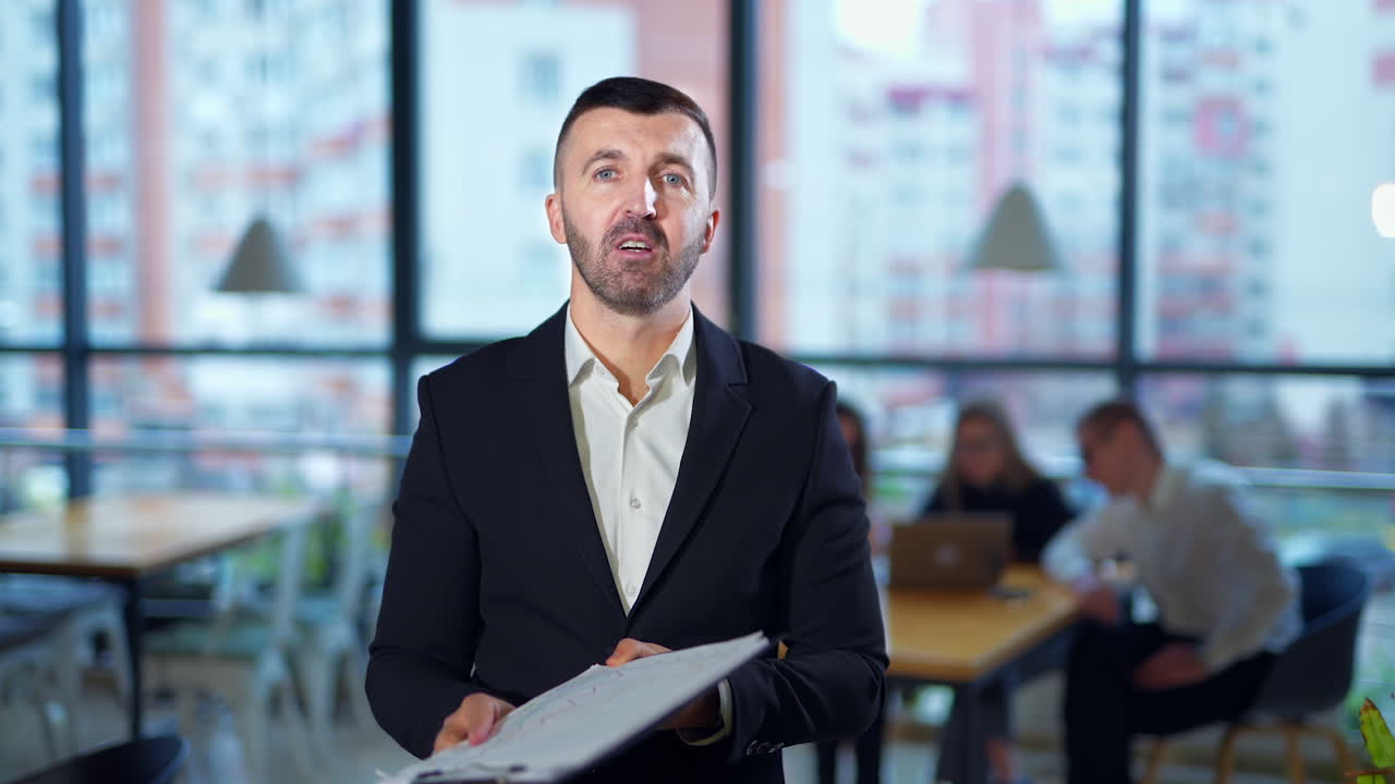 Serious man in suit talks in front of camera showing graphs. Confident male speaker performs presentation in office. People work at the desk at backdrop in blur.
