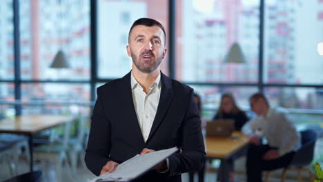 serious man in suit talks in front of camera showing graphs. confident male speaker performs presentation in office. people work at the desk at backdrop in blur.
