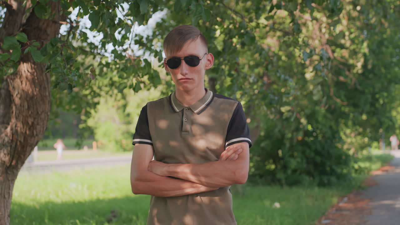 Man In Sunglasses Standing Under Tree Arms Crossed, Tense Guarded Stance In Leafy Park, Dappled Sunlight On Polo Shirt, Serious Expression Observing Surroundings, Solitary Watchful Mood