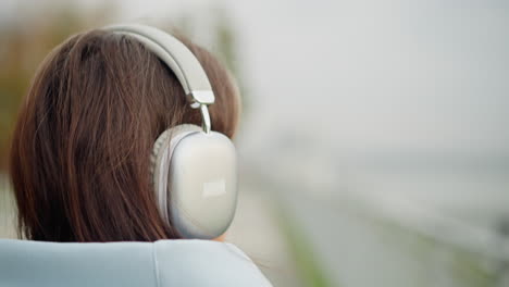 Rear view of student with brown hair wearing white headphones, strolling outdoors into blurred background, serene atmosphere, youthful and focused, peaceful walk in urban environment