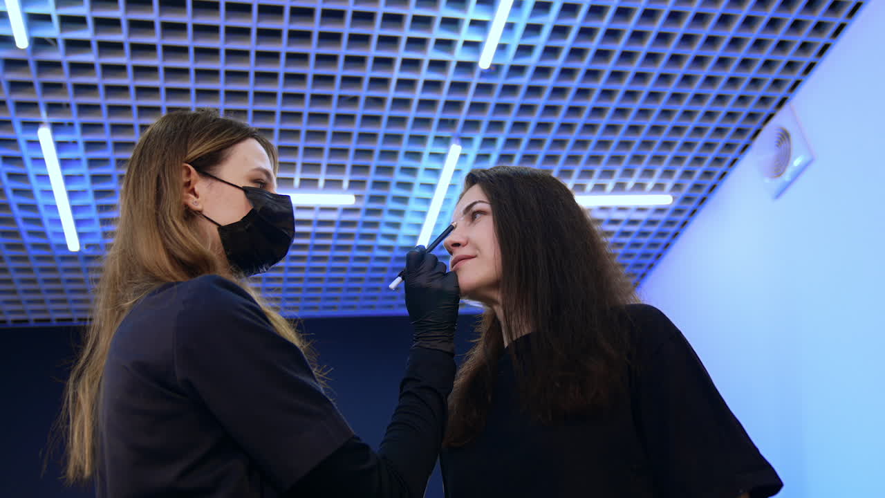 Beautician drawing white lines to mark the eyebrow limits. Brunette woman is prepared for the permanent eyebrow make up. Low angle view.