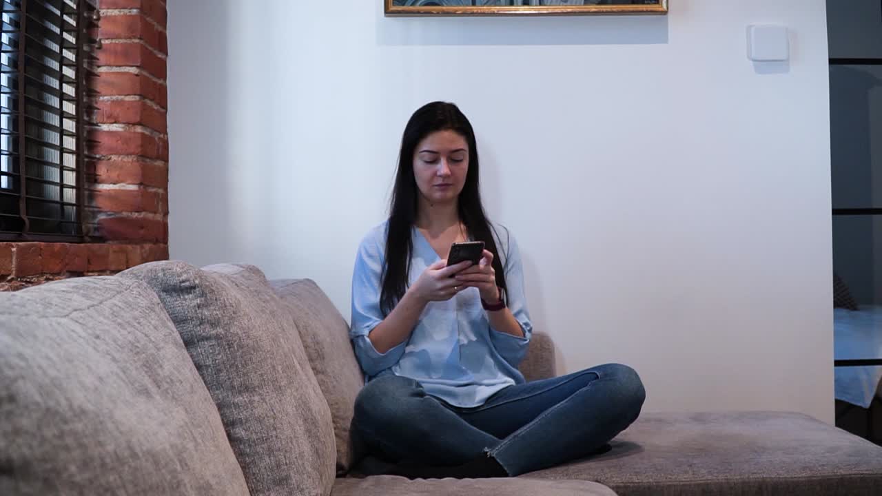 la joven está sentada en el sofá de la sala de estar con una pared de ladrillo rojo y está escribiendo un mensaje con su teléfono inteligente, tomado desde un lado