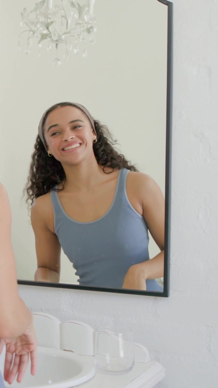 Video vertical: Frente al espejo, mujer con camiseta de tirantes azul aplicándose productos para el cuidado de la piel en el tocador, ajustándose una diadema