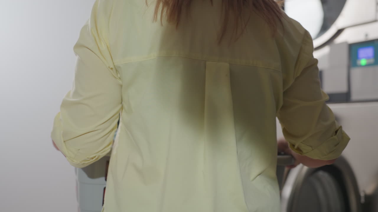Back view supervisor carrying laundry basket toward industrial washer row, door open, preparing to load garments inside laundromat, stainless panels visible, indoor service workflow