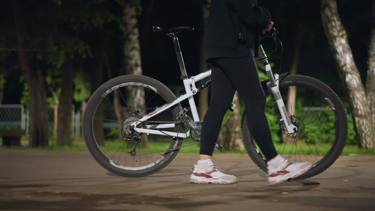 Nighttime City Stroll, Woman Walks Beside Illuminated Bike, Luminous Lamp Lights Up Quiet Night City Pavement Shadows, Woman In Sneakers Moves Slowly Past Glowing Street Lamps And Bicycles