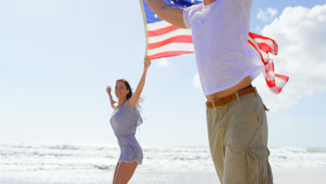 Vista-Lateral-De-Una-Joven-Pareja-Caucásica-Sosteniendo-La-Bandera-Estadounidense-Ondeando-En-La-Playa-4k