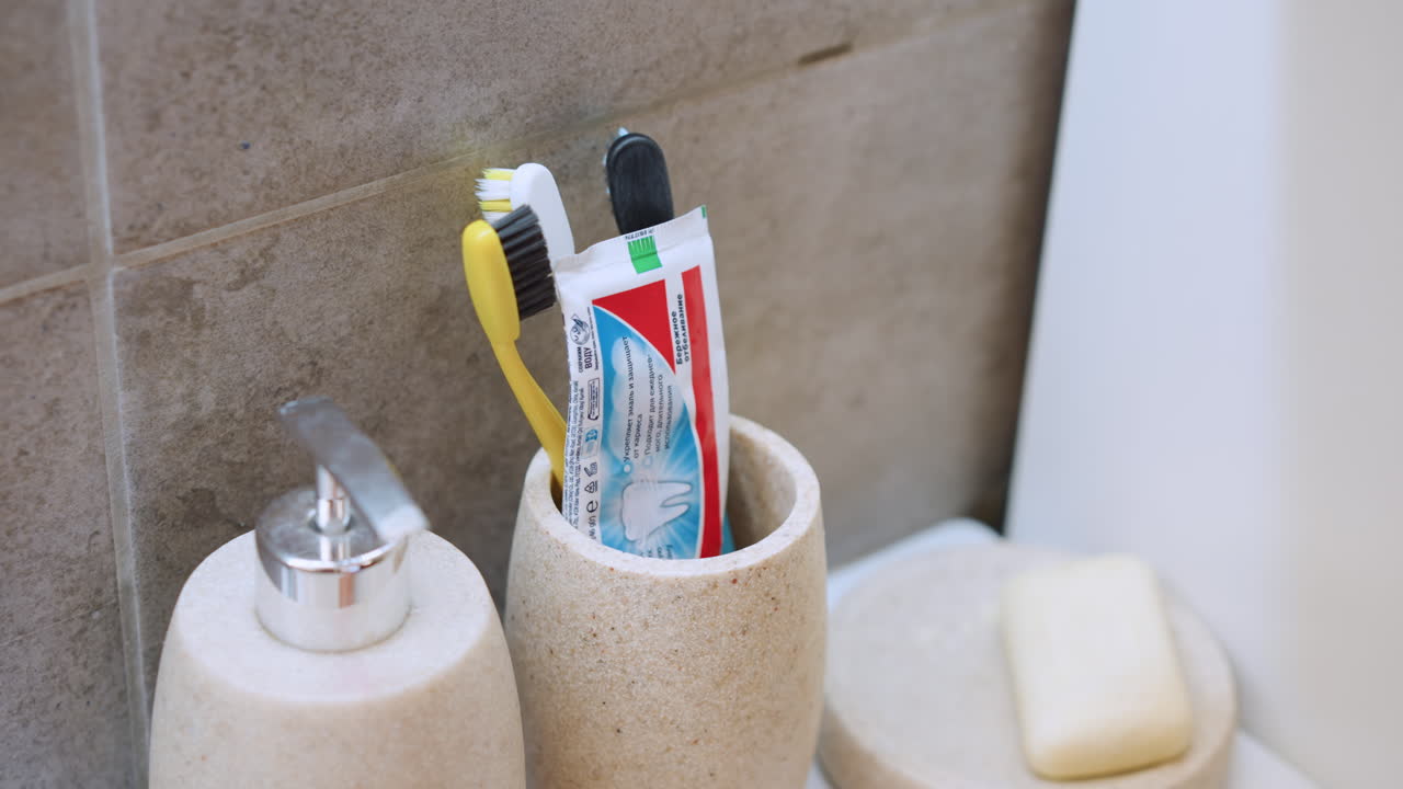 Ceramic cup filled with toothbrushes and tube of toothpaste on sink ledge, hand reaches to place green brush into holder, soap dish nearby, colorful bristles and neat arrangement, home bathroom