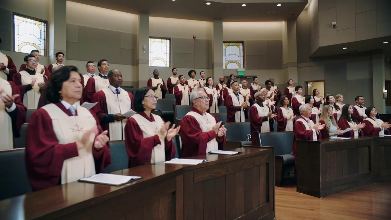 Coro aplaudiendo con túnicas granates y estolas blancas en los asientos de la iglesia, partituras de himnos en los atriles, la cámara haciendo un paneo.